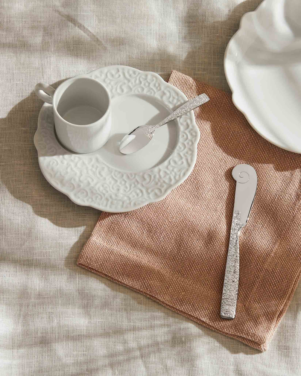 Embossed white porcelain and silver tableware by Alessi, including a coffee cup, saucer, spoon, and butter knife on a pink linen napkin.