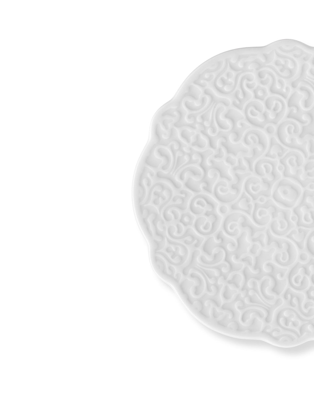 White ceramic plate by Alessi with a raised baroque pattern and scalloped edge, partially visible.