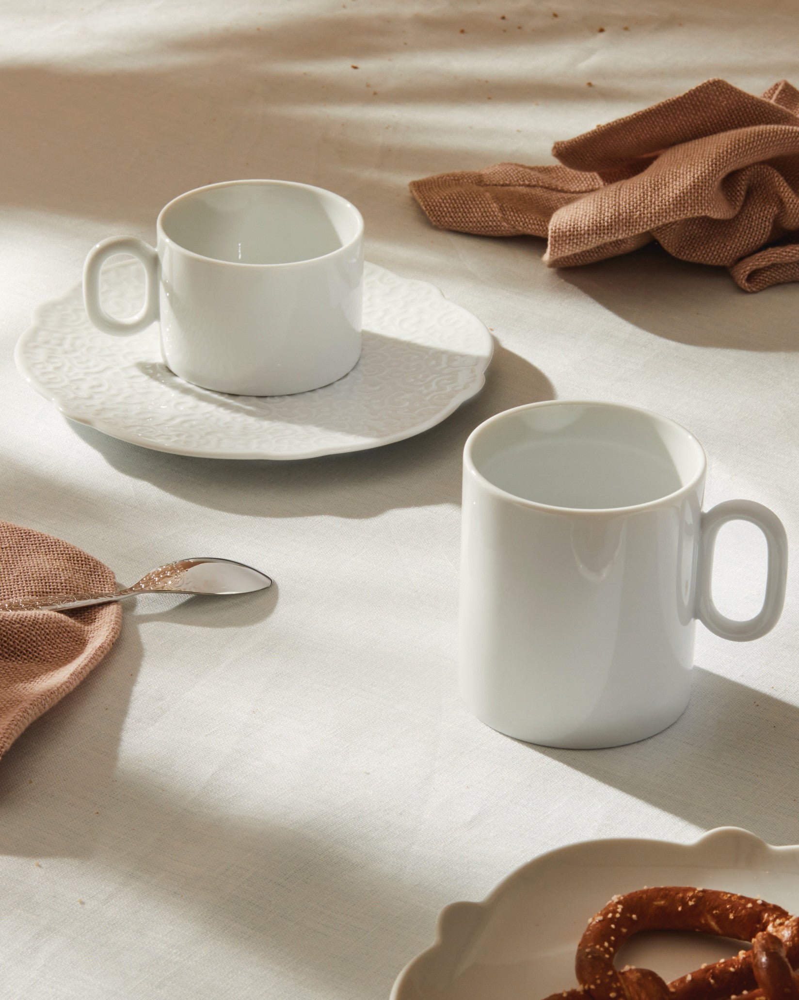 White ceramic mug by Alessi on an embossed saucer, styled with a second mug, a brown napkin, and pretzels.