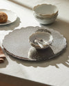 Silver platter by Alessi, with a scalloped edge and ornate embossed swirling pattern, displaying white bowls and a bread roll.