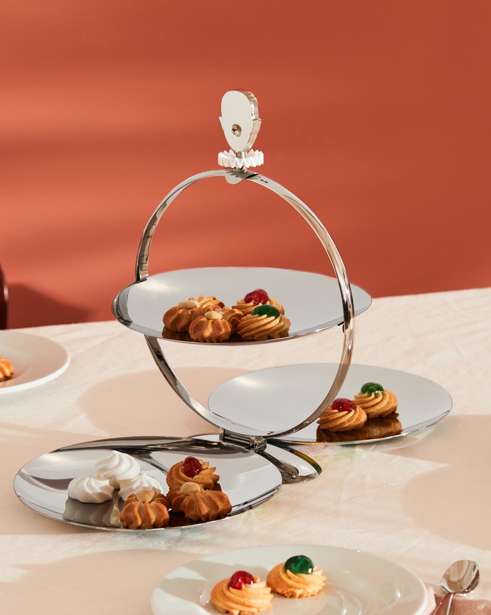 Silver folding tiered serving stand by Alessi, featuring a clown finial and various cookies.