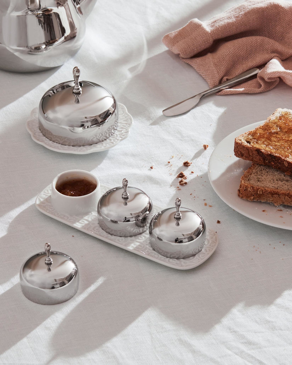 Silver lidded condiment dishes by Alessi with ornate white ceramic bases on a breakfast table.