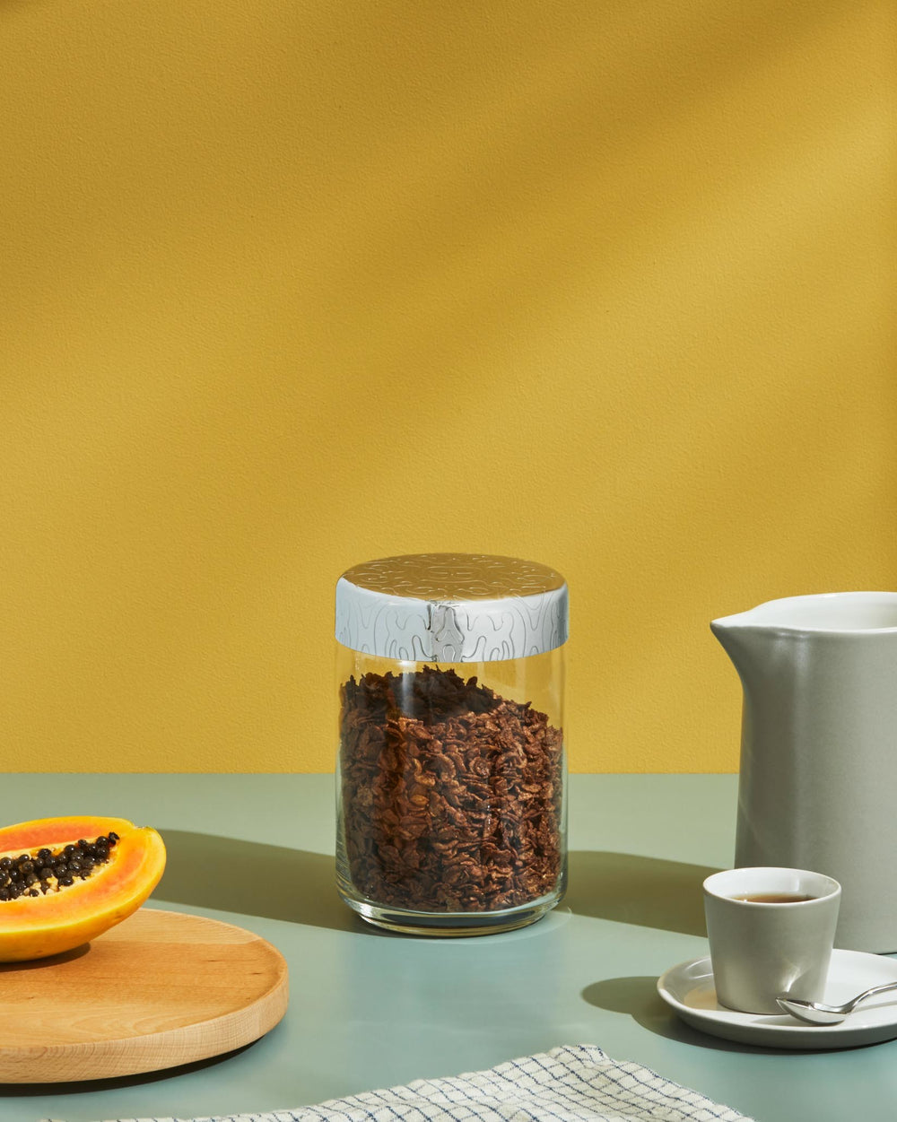 Clear glass storage jar by Alessi with a decorative silver patterned lid, holding dark granola.