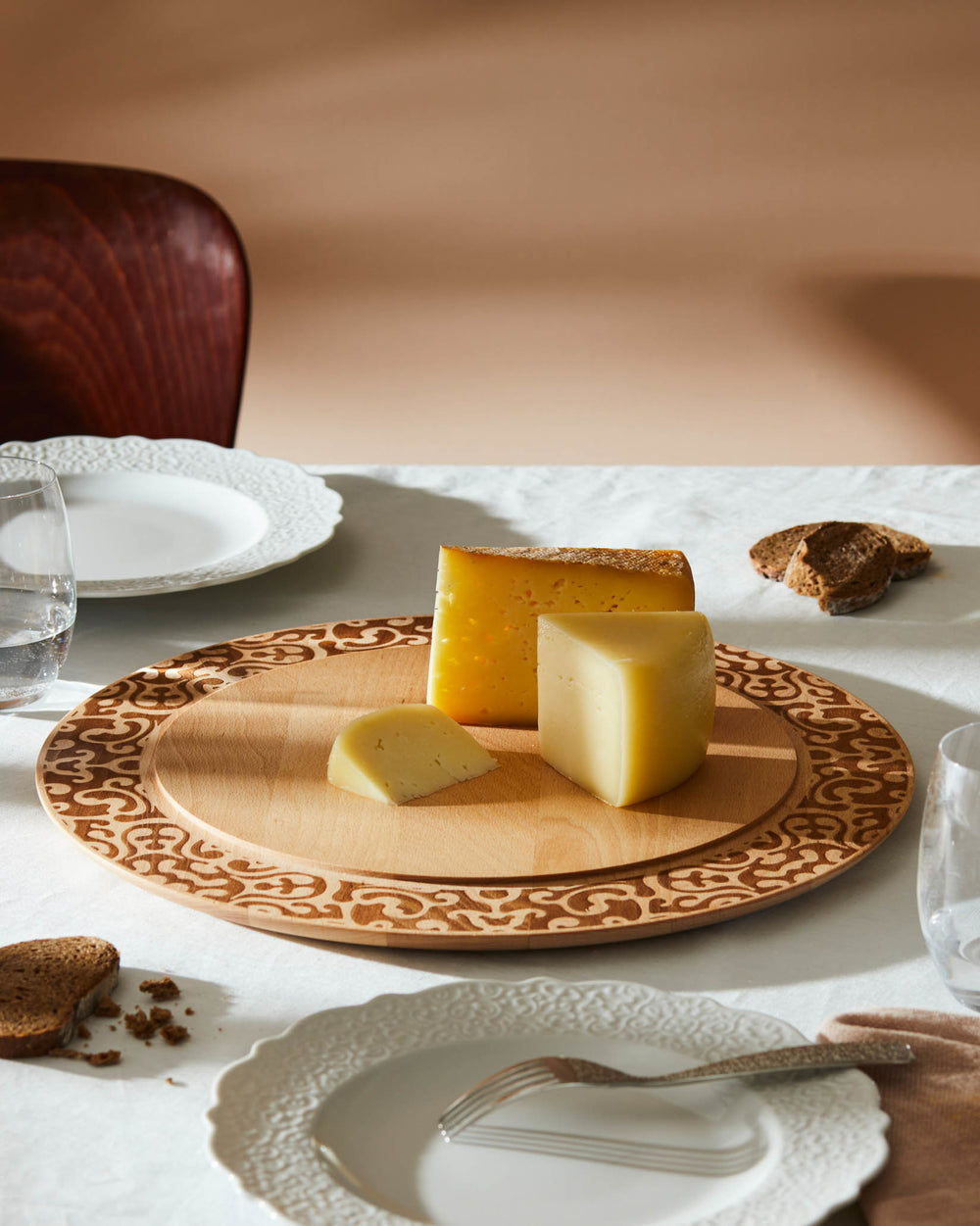 Round wood chopping board by Alessi with an engraved patterned edge, styled with cheese and bread.