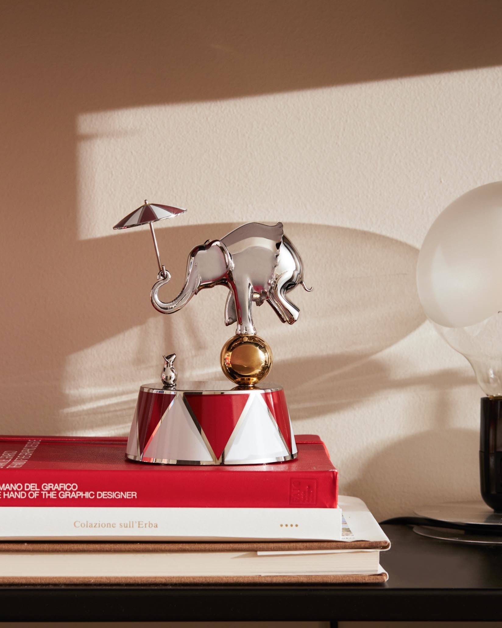 Chrome-plated elephant figurine by Alessi, balancing on a golden ball atop a red and white striped base, with a tiny mouse and umbrella.
