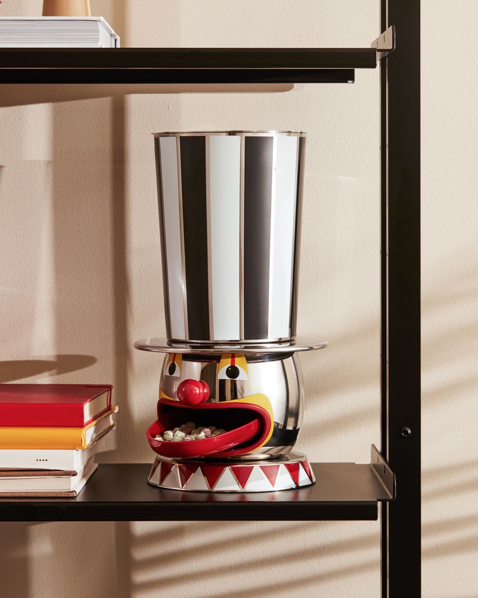 Clown-shaped candy dispenser by Alessi, featuring a black and white striped top hat and a red mouth filled with candies.