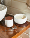 Silver honey dipper by Alessi, with a polished spherical handle and flat tip, on a wooden countertop next to a white honey pot.