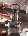 Polished stainless steel teapot by Alessi with black handle and lid knob, surrounded by matching serving pieces and breakfast items.