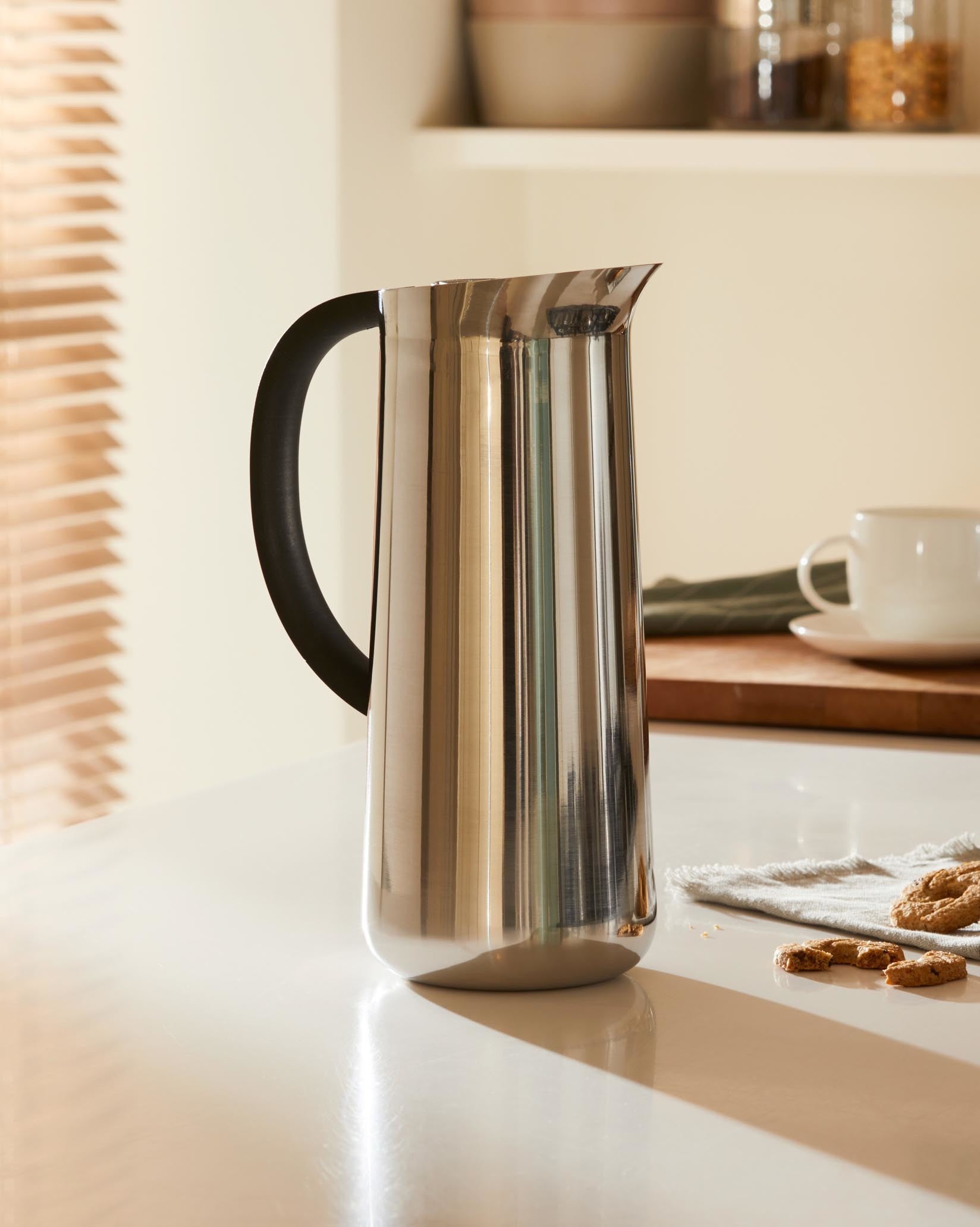 Tall, sleek stainless steel pitcher by Alessi with a black handle, standing on a reflective light countertop.
