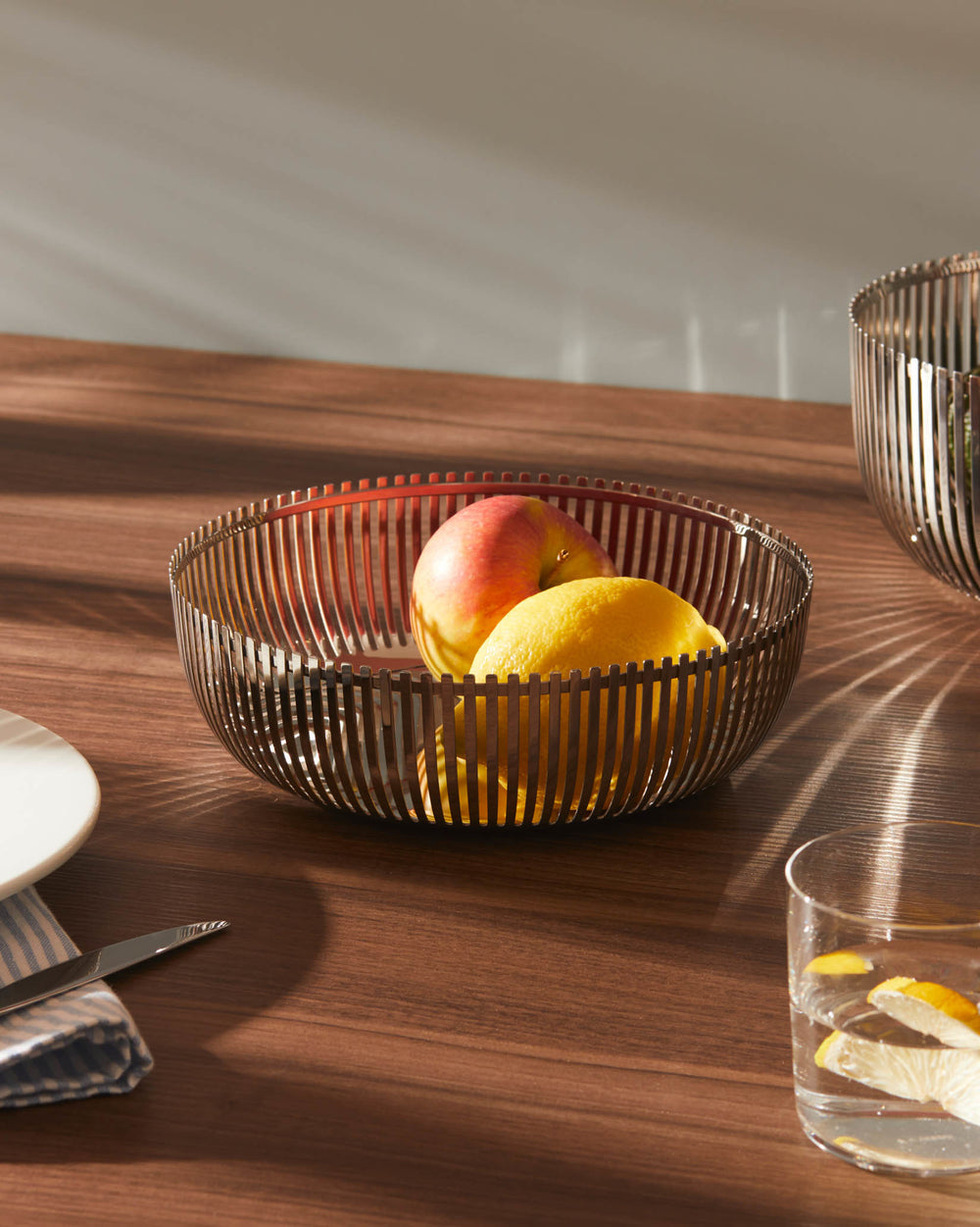 Bronze slatted fruit bowl by Alessi, holding an apple and lemons on a warm wooden table.