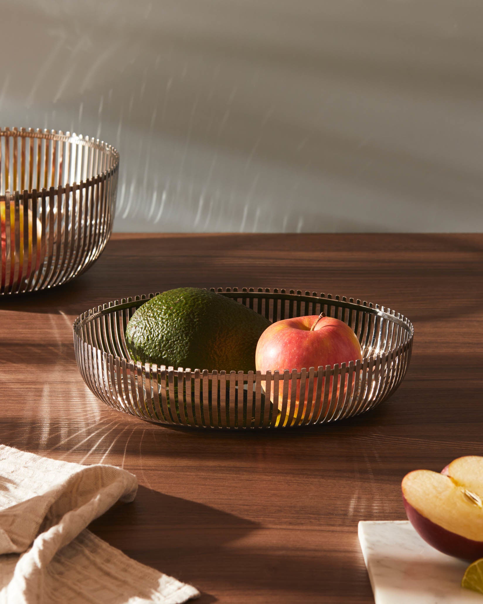Metallic slatted fruit basket by Alessi filled with an avocado and a red apple.