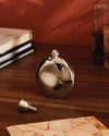 Shiny metal oval flask by Alessi, with screw cap and funnel, on a wooden table.