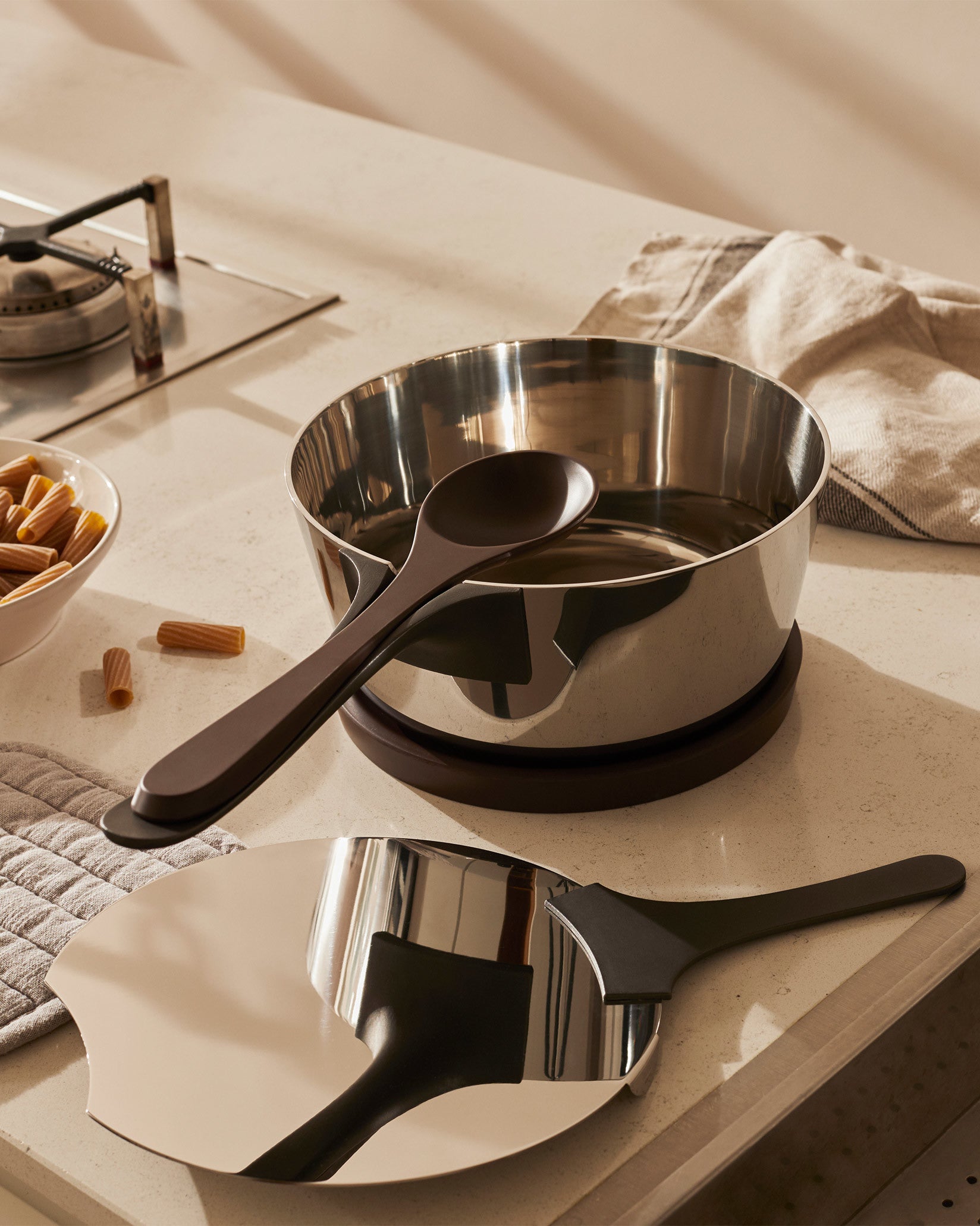 Polished stainless steel pasta pot by Alessi with a dark brown serving spoon and matching cooking tool, on a kitchen counter.