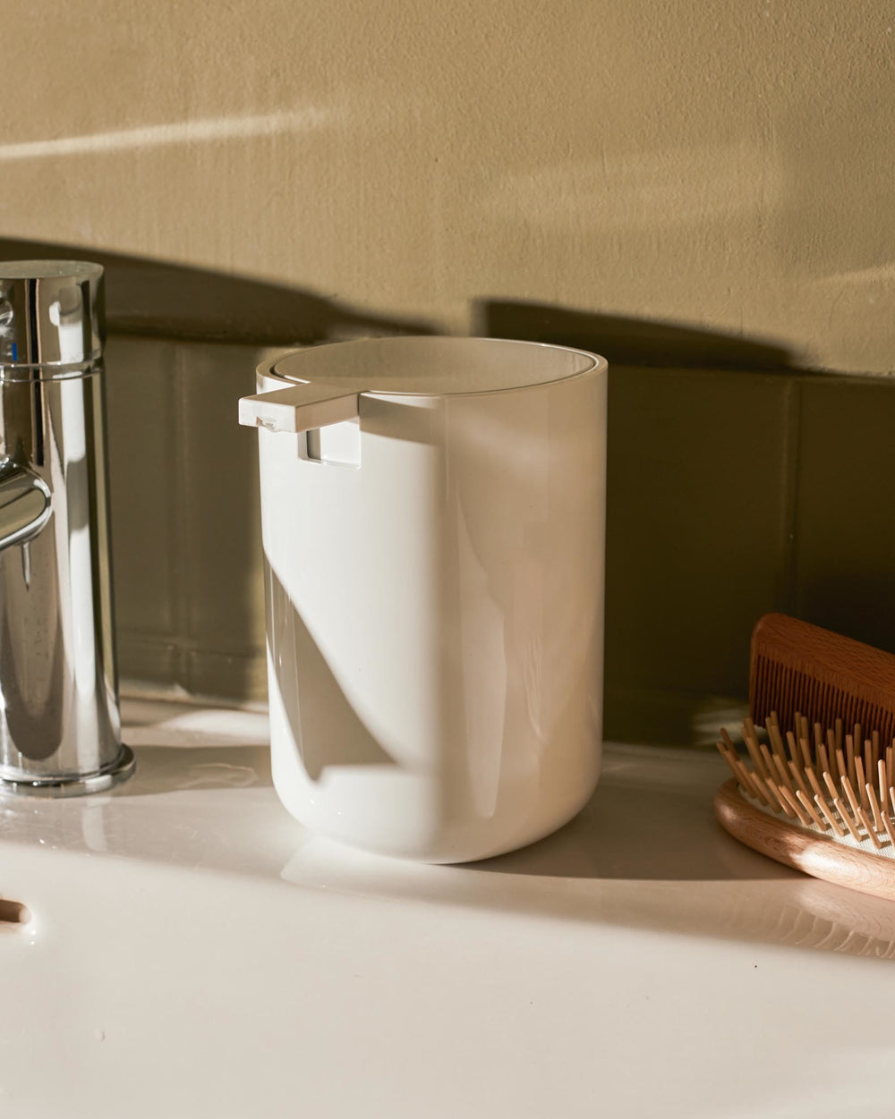 White cylindrical dispenser by Alessi with a flat top on a white bathroom counter, beside a chrome faucet and wooden brush.