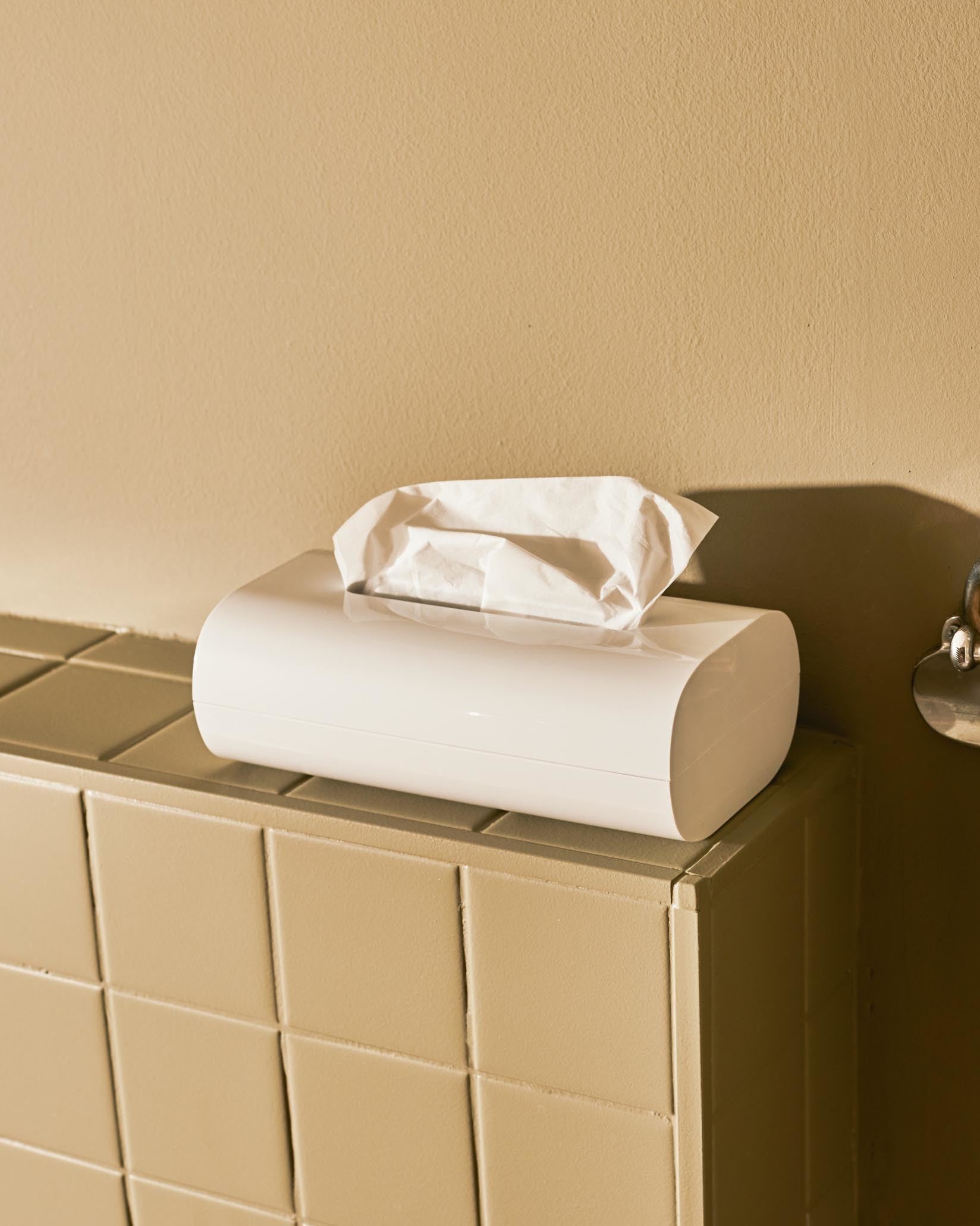 White tissue box holder by Alessi with curved ends and a white tissue emerging, on a beige tiled surface.