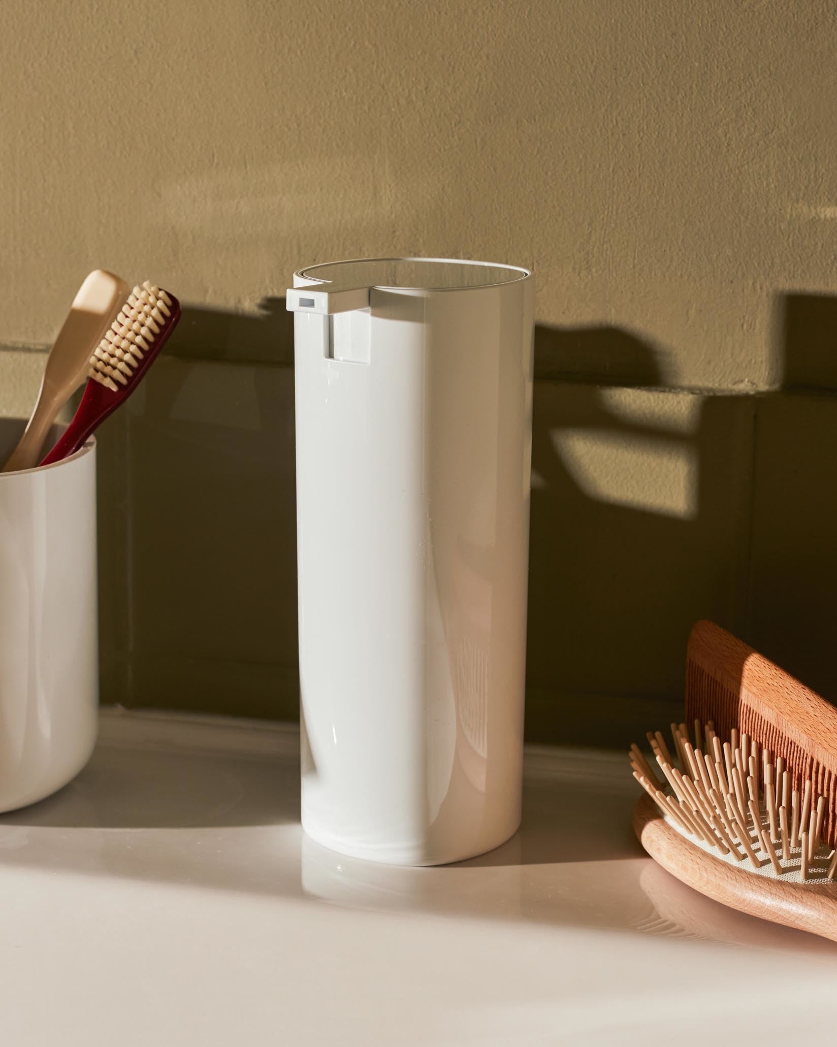 White soap dispenser by Alessi, on a light countertop next to toothbrushes and a wooden hairbrush.