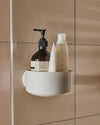 White wall-mounted shower caddy by Alessi, holding three assorted bottles on a beige tiled wall.
