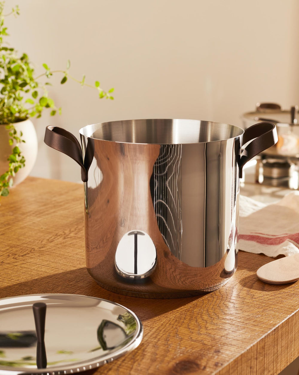 Polished stainless steel cooking pot by Alessi, featuring dark brown loop handles and a circular design element, with its lid on a wooden counter.