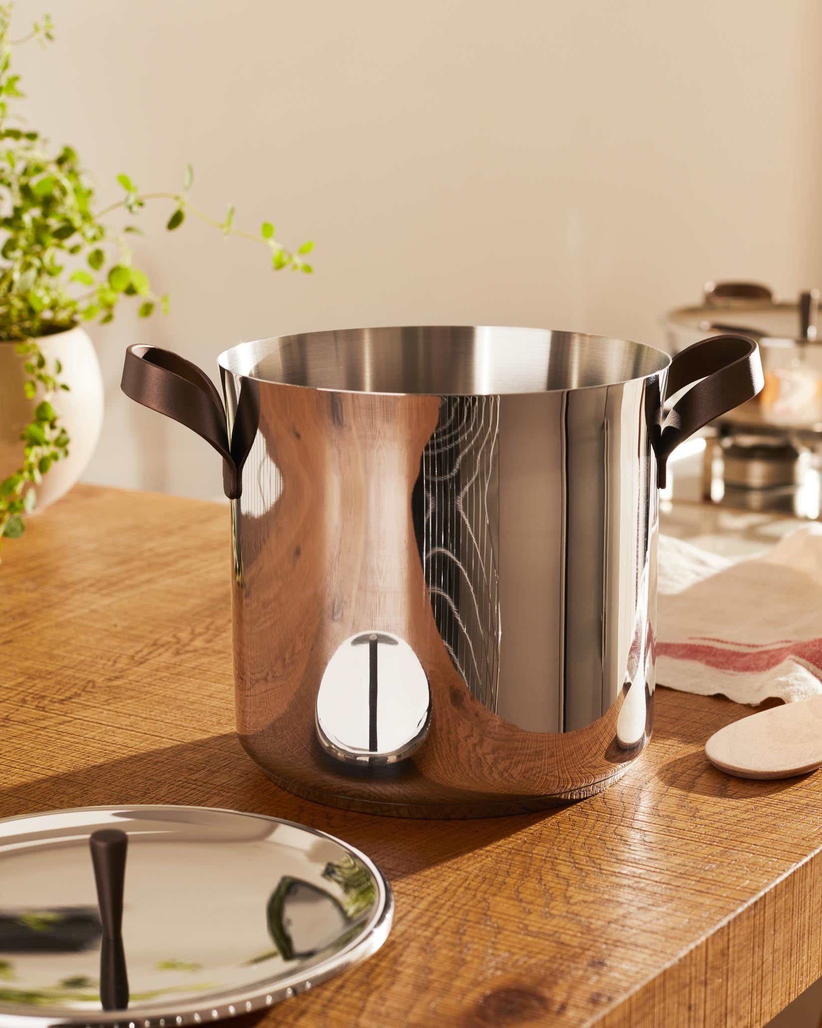 Polished stainless steel cooking pot by Alessi, featuring dark brown loop handles and a circular design element, with its lid on a wooden counter.