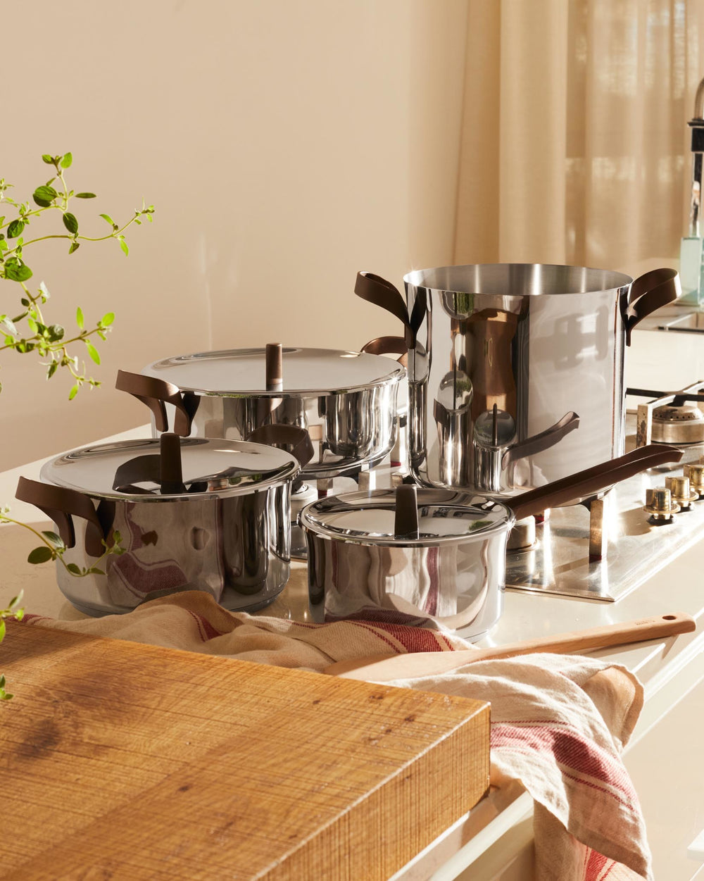 Stainless steel pots with brown handles by Alessi, a set of four displayed on a kitchen counter with a wooden board.