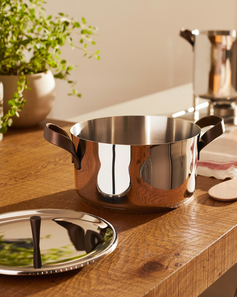 Stainless steel casserole pot by Alessi with brown leather handles on a rustic wooden table, lid nearby.
