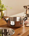 Polished stainless steel casserole with brown handles by Alessi on a wooden countertop with a matching lid.