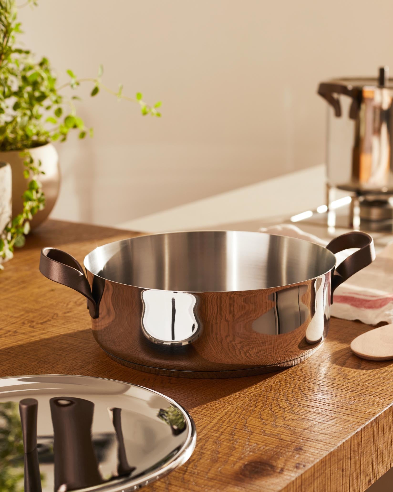 Polished stainless steel casserole with brown handles by Alessi on a wooden countertop with a matching lid.