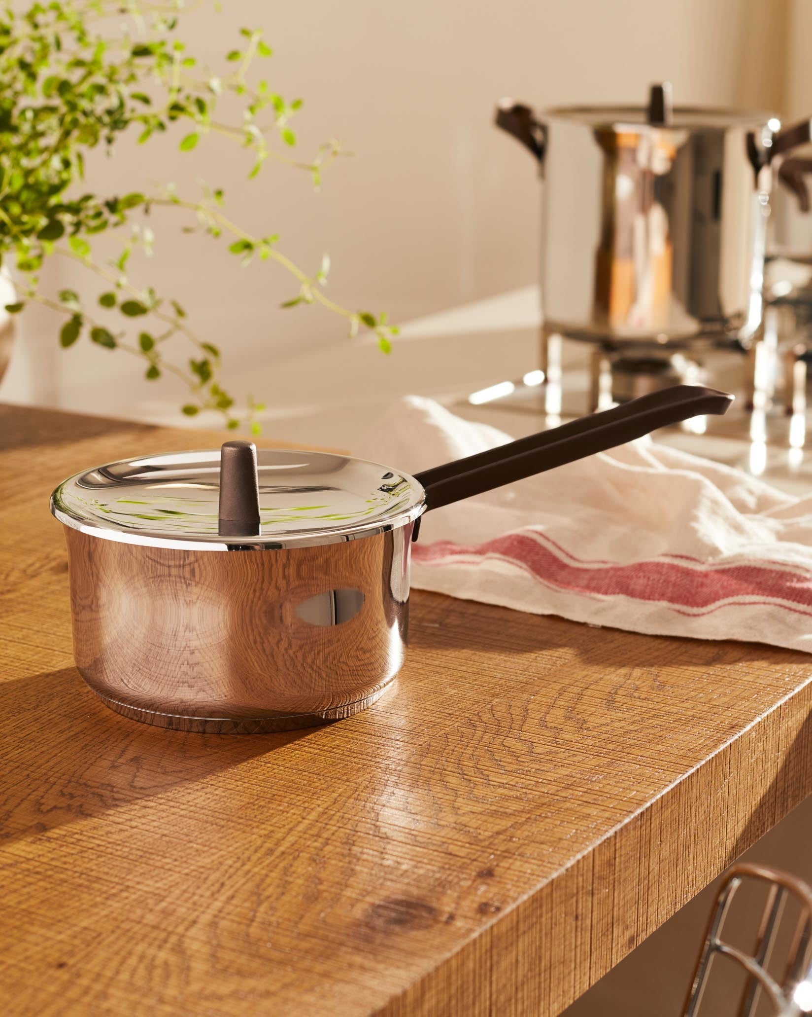 Stainless steel saucepan by Alessi with a dark brown handle and lid knob, on a wooden counter with a striped tea towel.