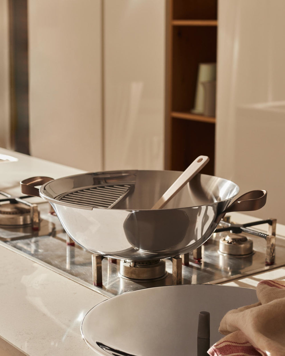 Stainless steel wok pot by Alessi with copper-colored handles, containing a metal rack and wooden spoon on a gas stovetop.