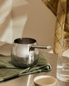 Silver metallic saucepan with a rounded base by Alessi, featuring a short handle and pour spout, resting on a green striped tea towel.
