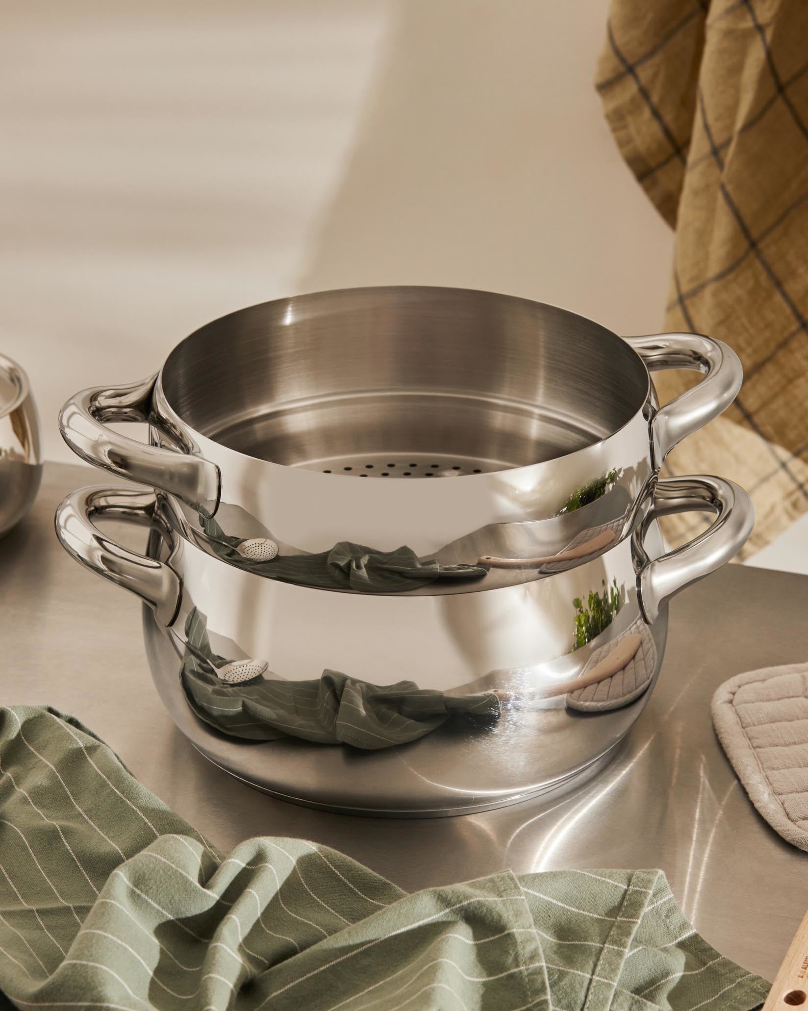 Two-tiered Mami stainless steel steaming pots by Alessi, stacked on a metal surface, with green and plaid kitchen towels.