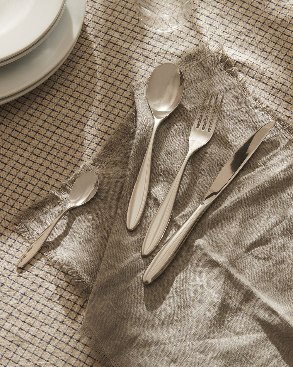 Polished silver four-piece cutlery set by Alessi, laid on a wrinkled grey linen napkin and beige checkered tablecloth.
