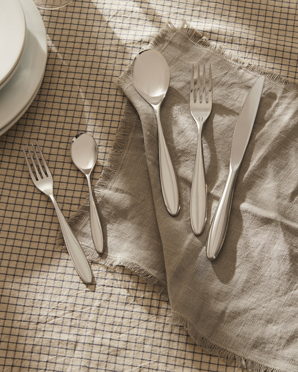 Mami silver cutlery set by Alessi on a beige plaid tablecloth with a fringed napkin.