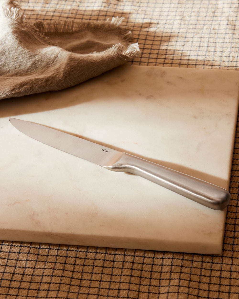 Silver metallic knife by Alessi, with an ergonomic handle, angled on a light marble cutting board and a brown checkered cloth with frayed edges.