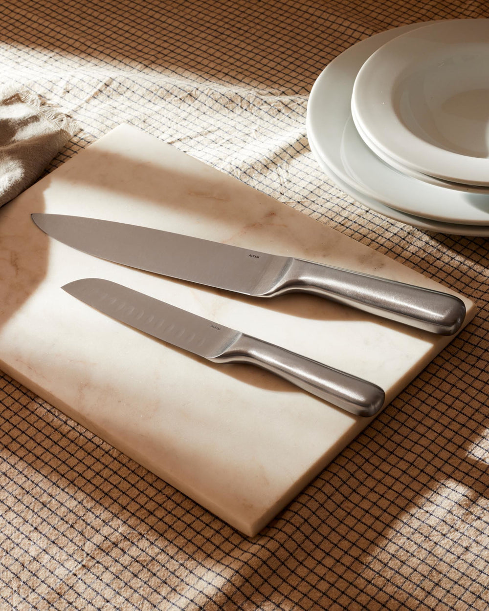 Stainless steel knives by Alessi on a marble board, with a checkered tablecloth and stacked white plates.
