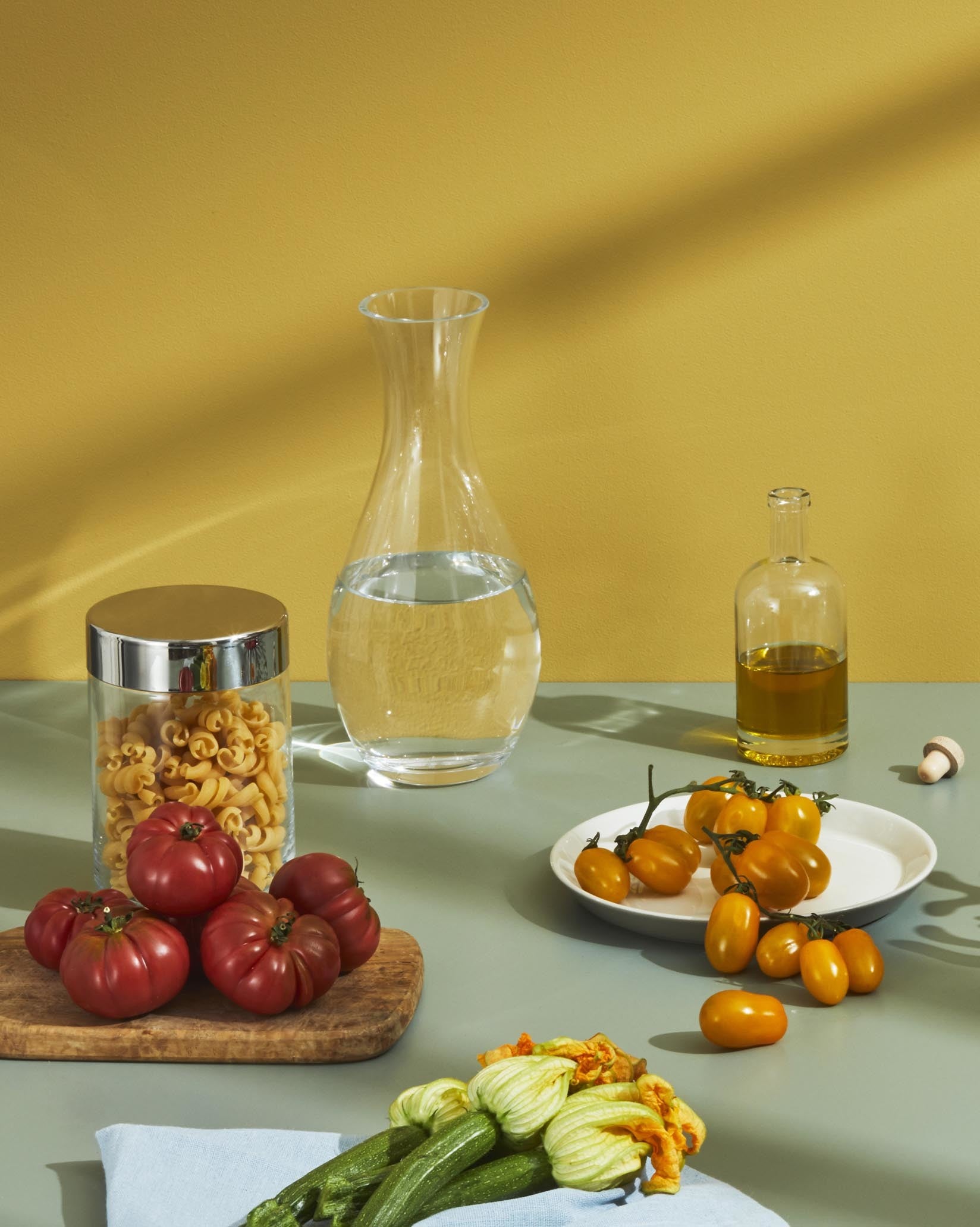 Elegant clear glass water carafe by Alessi, filled with water, styled on a table with fresh ingredients.