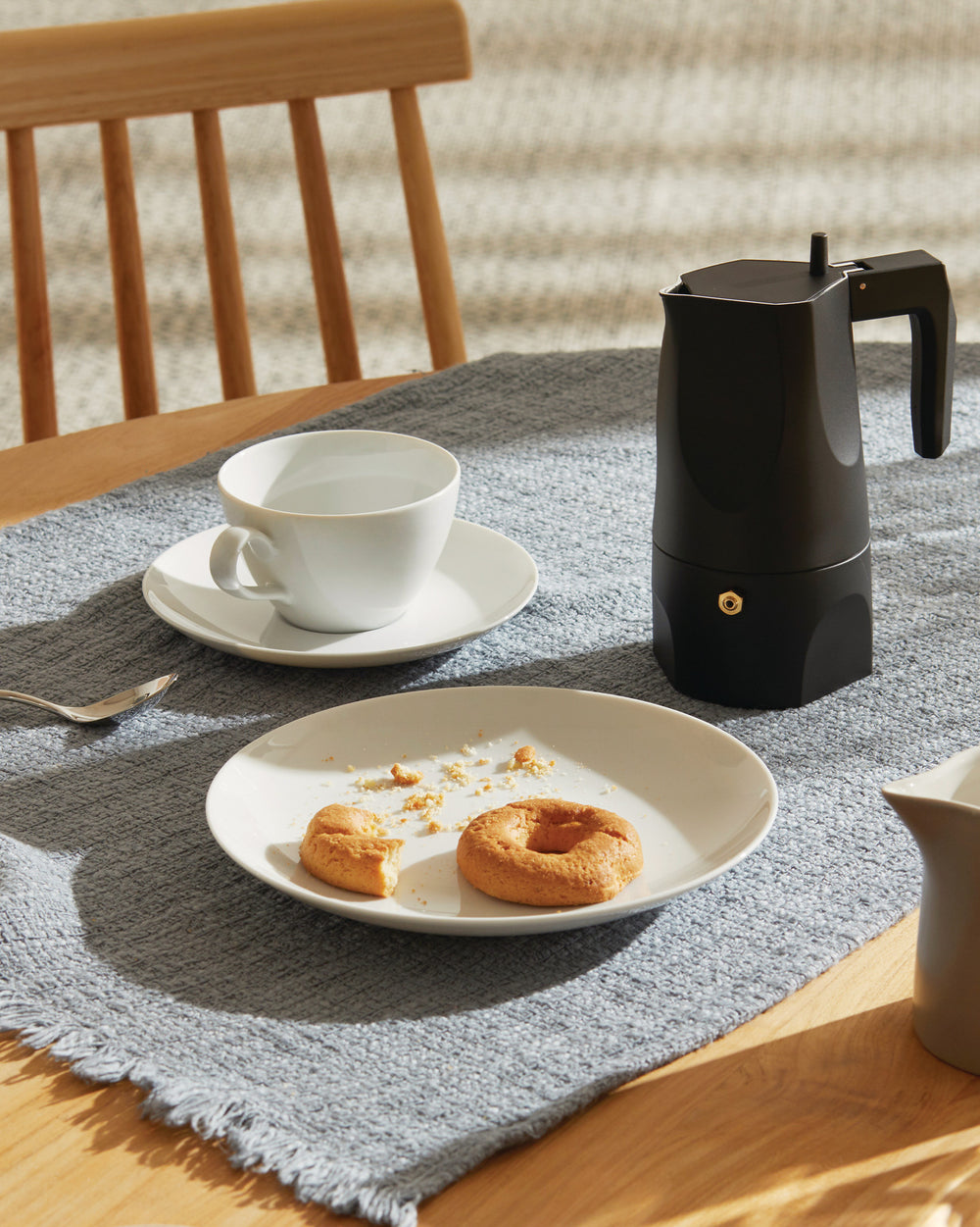 Black modern espresso maker by Alessi with a gold valve, on a grey placemat beside cookies and a white cup.