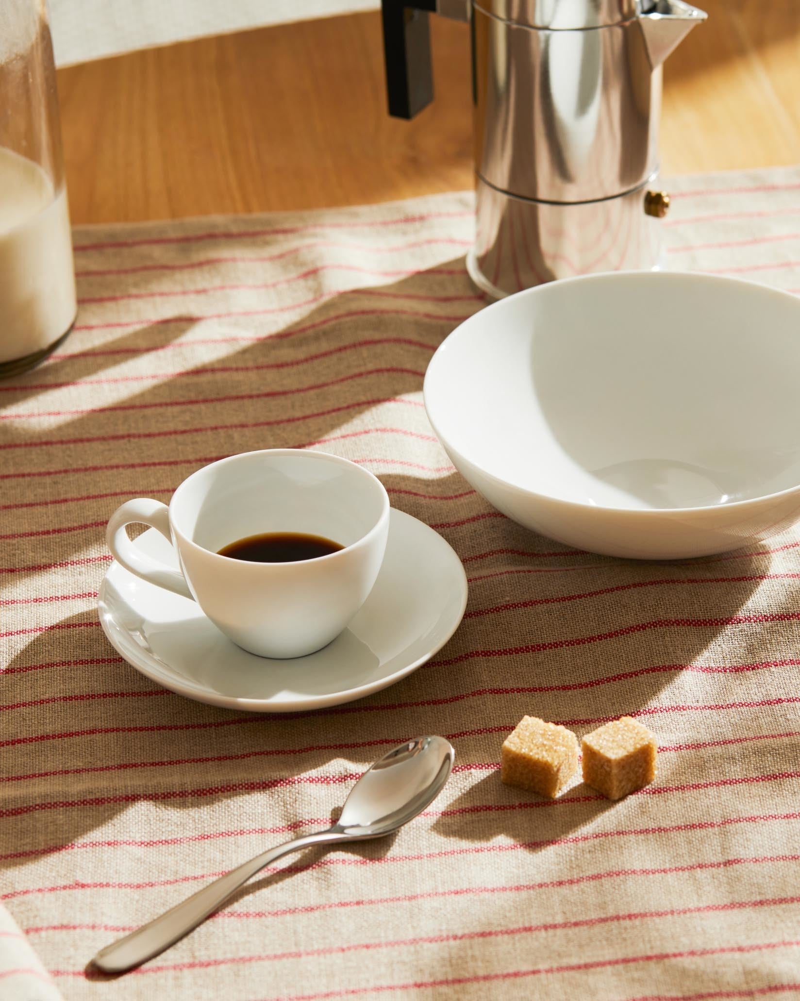 White ceramic coffee cup by Alessi, with a matching saucer, large bowl, moka pot, spoon, and sugar cubes on a striped tablecloth.