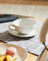 White ceramic cup and saucer by Alessi on a grey striped placemat, next to a plate with a pink macaron and yellow dessert pieces.