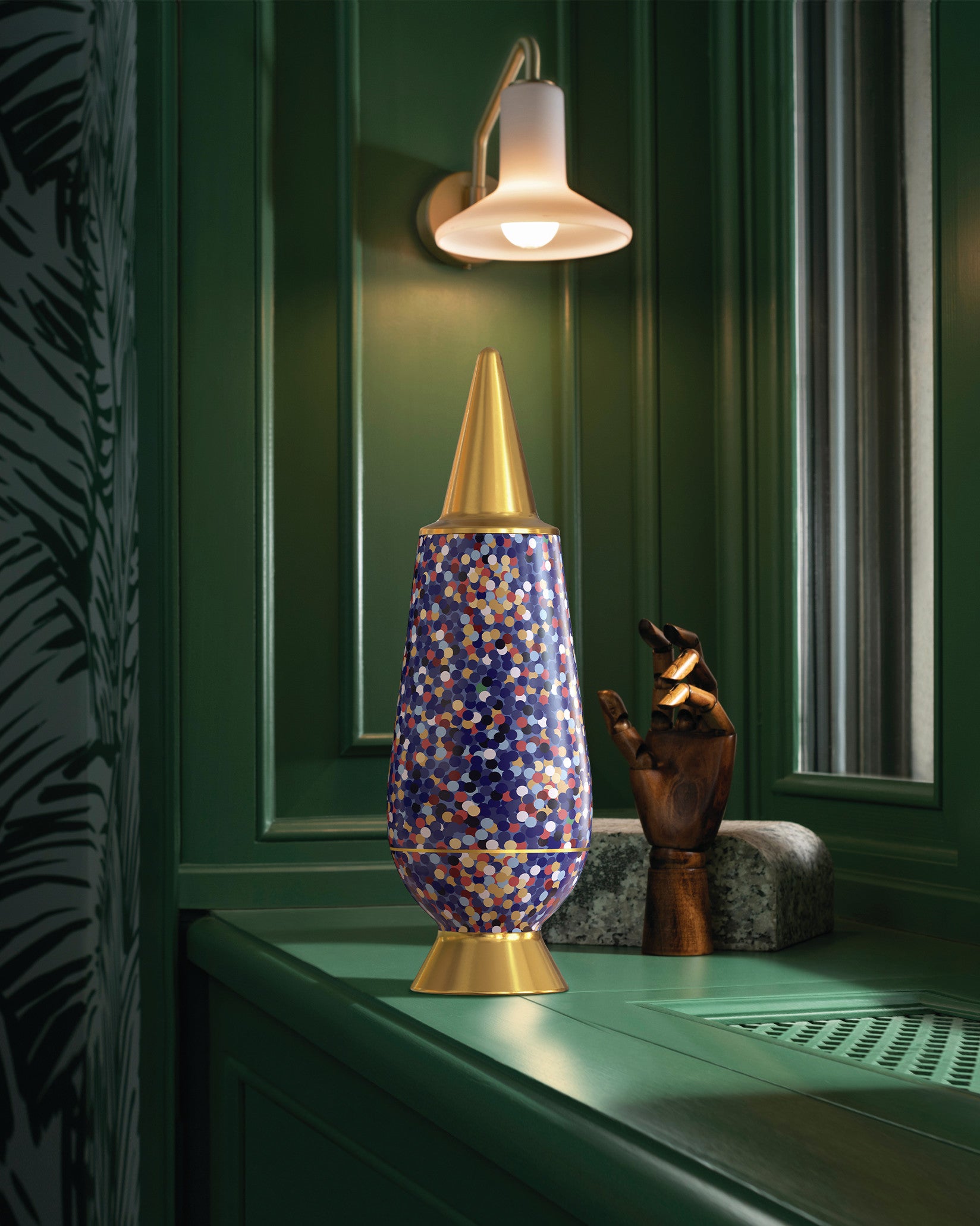 Multi-colored confetti patterned decorative item with gold accents by Alessi, standing on a dark green counter.