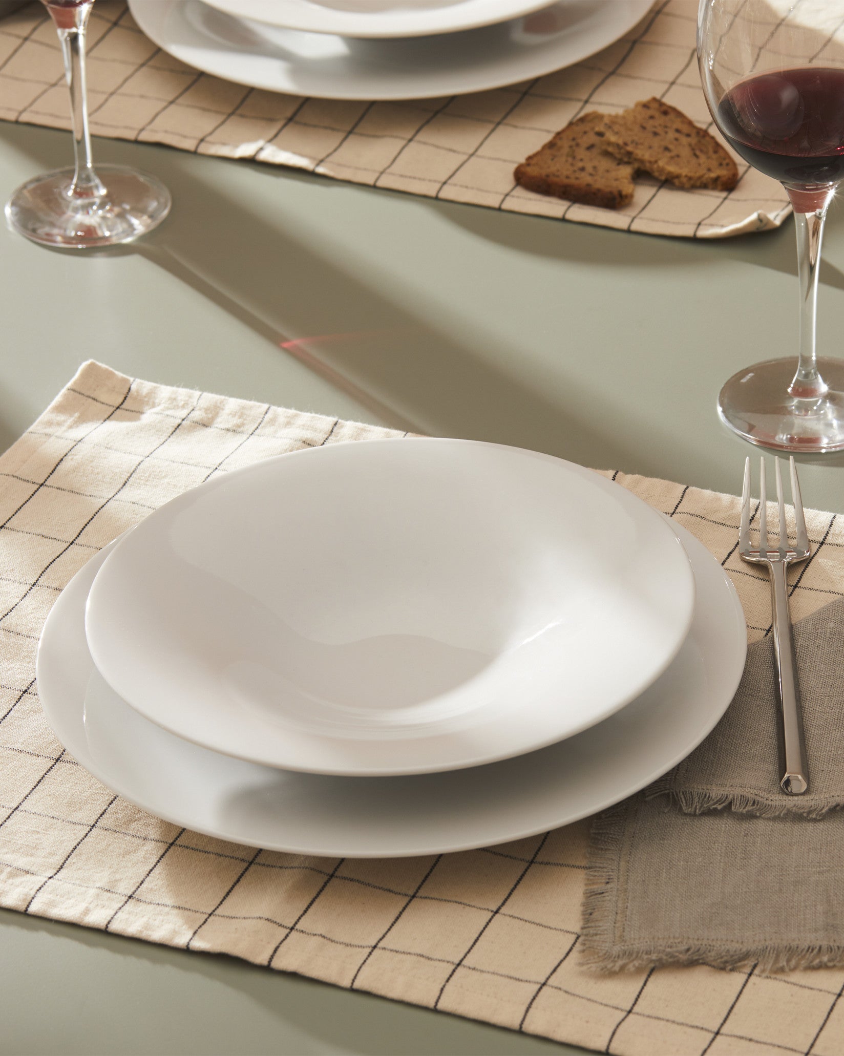 White dinnerware set by Alessi, stacked on a beige checkered placemat with a fork and wine glasses.