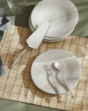 Silver serving cutlery by Alessi, including a cake server, fork, and spoon, styled on a pale grey plate over a beige checked tablecloth.