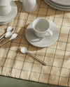 White ceramic teacup and saucer by Alessi, displayed on a beige plaid tablecloth with spoons and other white dishes.