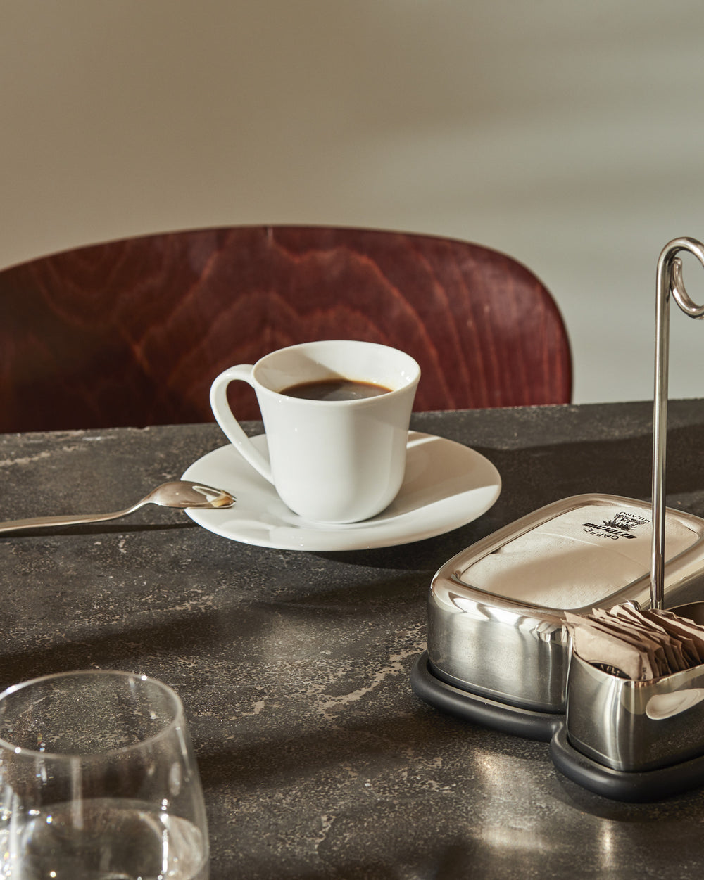 Sleek, modern white porcelain coffee cup by Alessi on a dark textured table with a spoon, napkin dispenser, and water glass.
