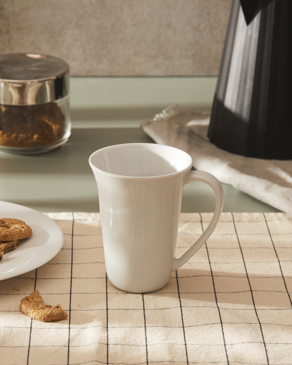 White ceramic mug with flared rim by Alessi, displaying subtle crackle texture on a checkered tablecloth.