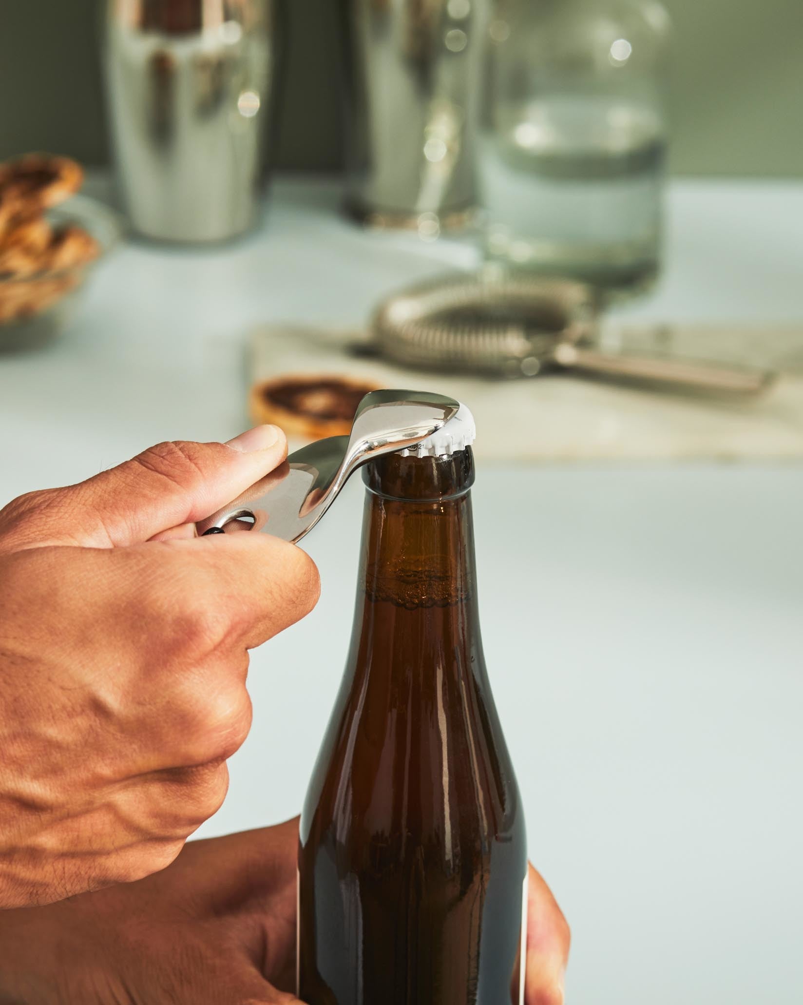 Silver metallic bottle opener by Alessi opening a brown beer bottle held by a hand.