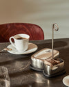 Polished stainless steel table organizer by Alessi, featuring a napkin holder, sugar packet compartment, and a vertical loop, on a textured dark table.