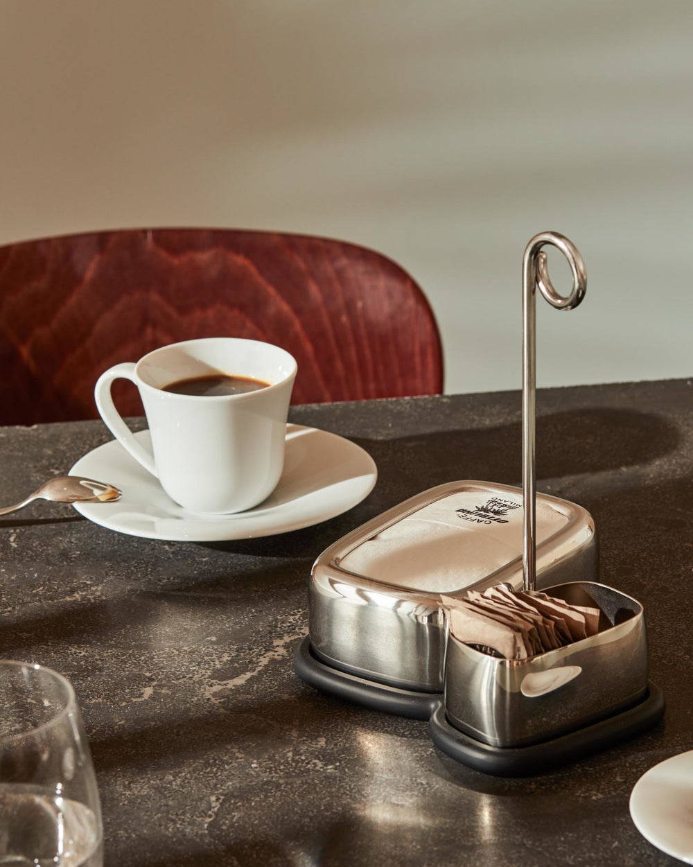 Polished stainless steel table organizer by Alessi, featuring a napkin holder, sugar packet compartment, and a vertical loop, on a textured dark table.