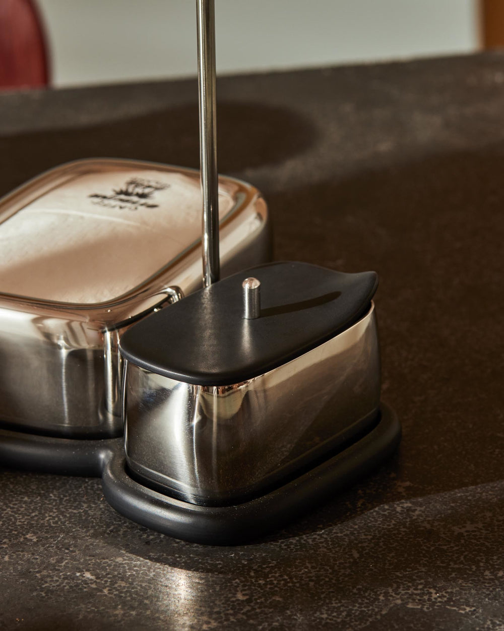 Polished stainless steel condiment set by Alessi, with black lids and a unifying black base on a dark textured surface.