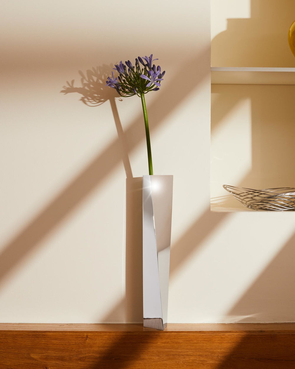A polished silver Crevasse vase by Alessi, featuring an angular geometric design, holding a single purple flower.